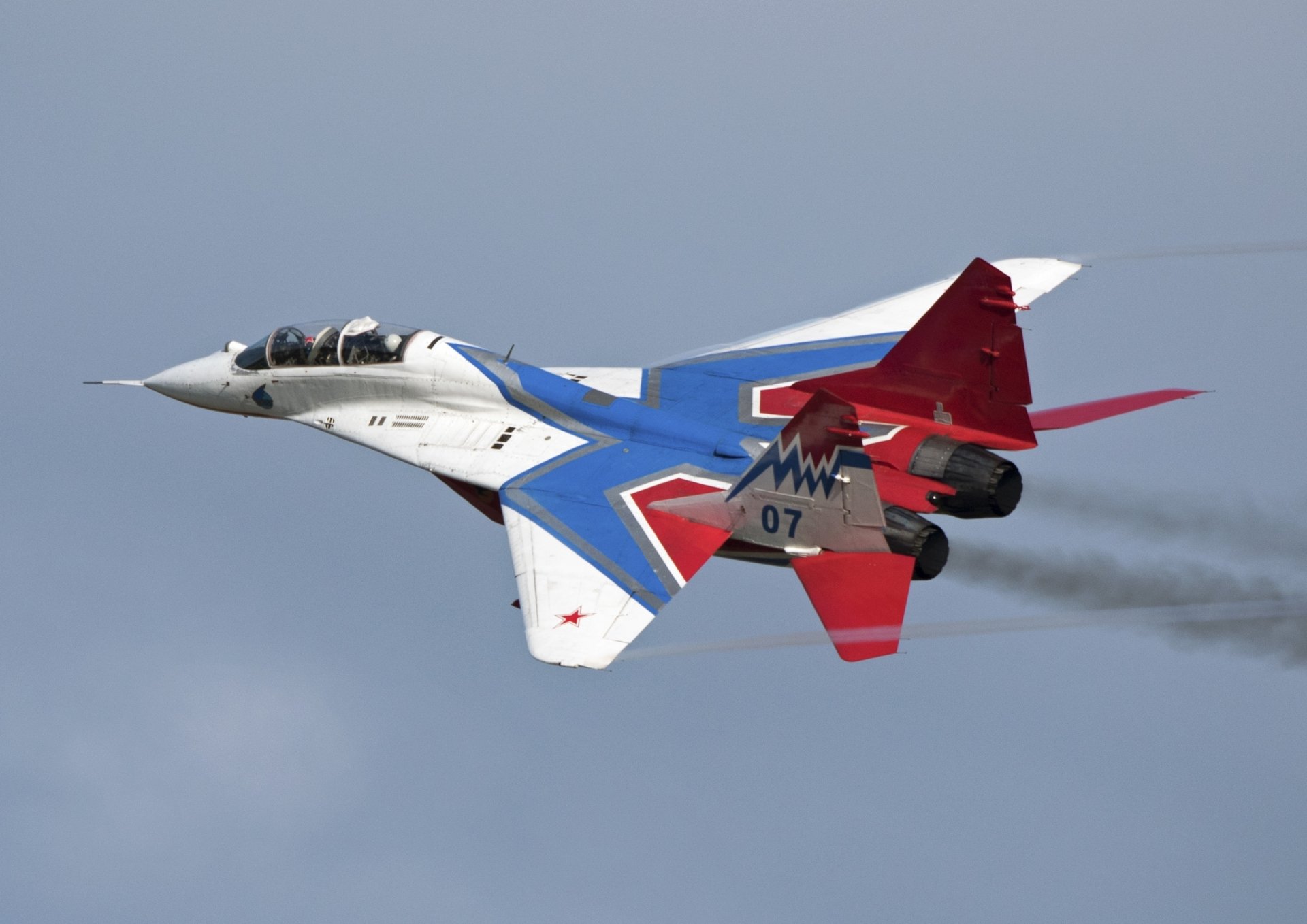 Military Mikoyan MiG-29 jet fighter warplane in red, white and blue livery trailing smoke over a cloudy sky — 2K Quad HD PC desktop wallpaper background.