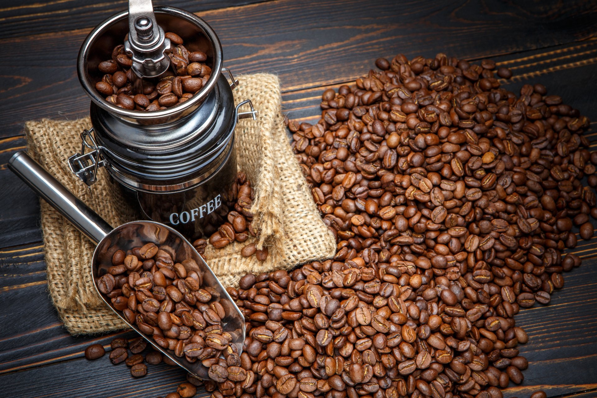 Close-up of roasted coffee beans spilling from a metal scoop beside a vintage coffee grinder on a dark wooden surface, captured in 4K Ultra HD for a PC desktop wallpaper.