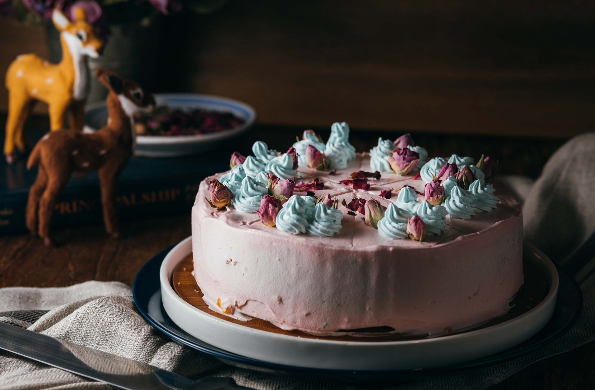 HD desktop wallpaper of a cream-covered pastry cake dessert topped with pastel blue and pink whipped-rosettes and dried rose petals on a plate against a dark wooden background.