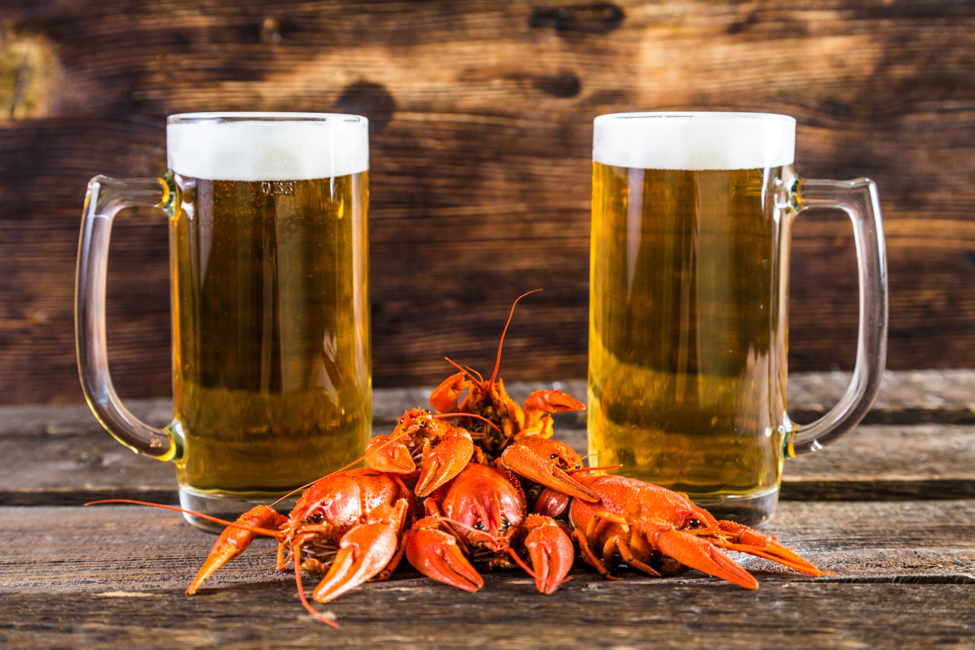 Two frosty beer mugs flank a pile of bright red crayfish on a rustic wooden surface, captured in vivid 4K Ultra HD for a rich food and drink desktop background.