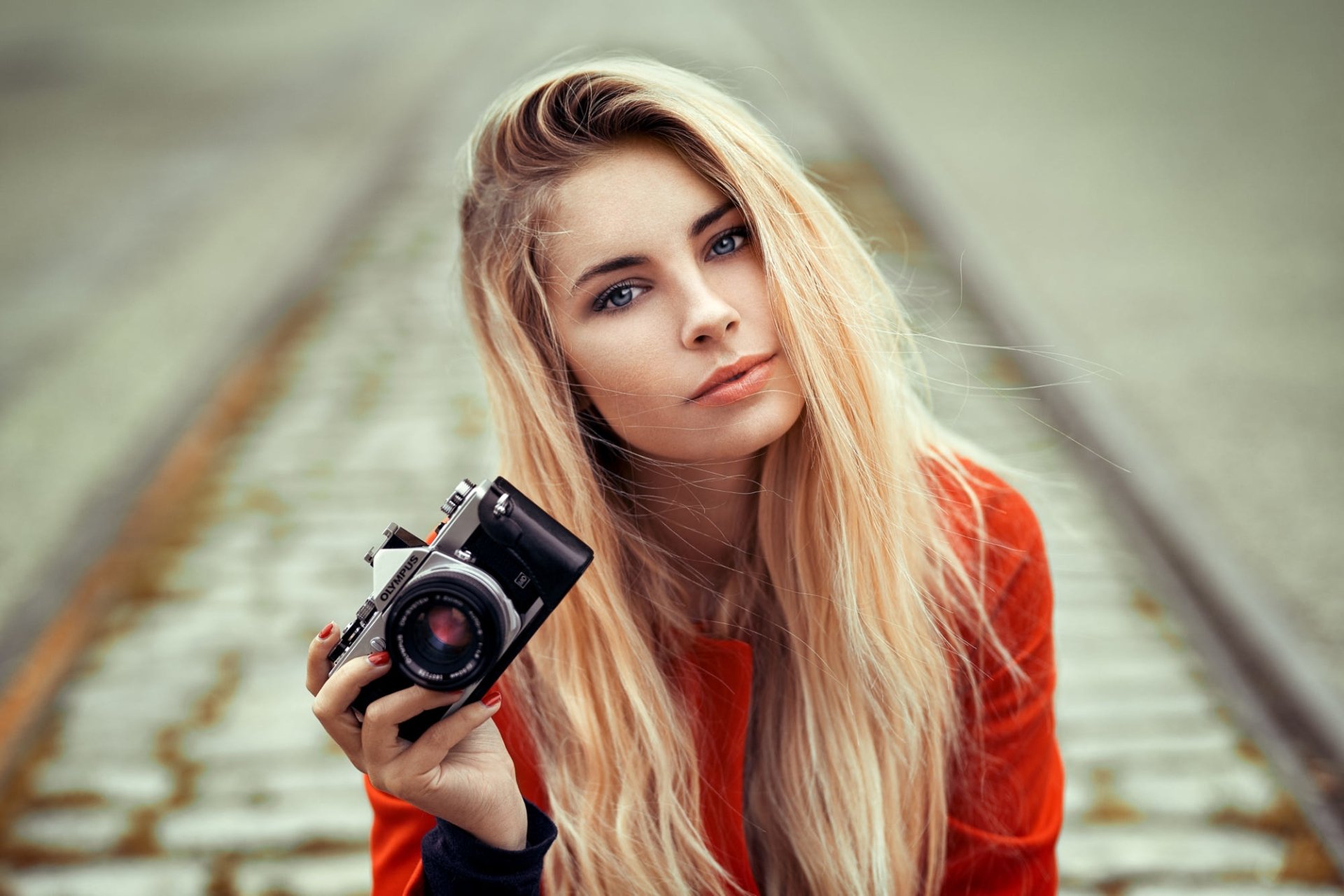 A blonde woman model with blue eyes holding an Olympus camera, captured with shallow depth of field in an HD desktop wallpaper.