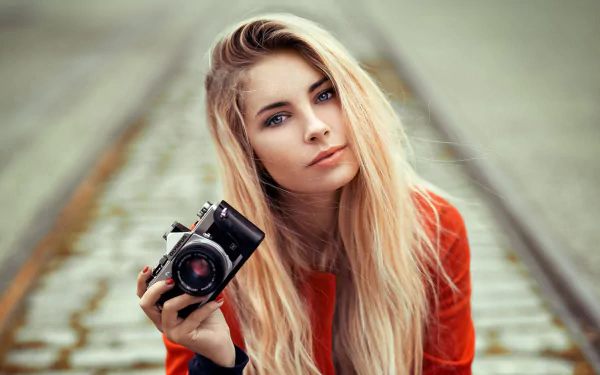 A blonde woman model with blue eyes holding an Olympus camera, captured with shallow depth of field in an HD desktop wallpaper.