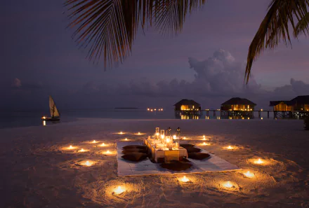 4K Ultra HD PC desktop wallpaper: candlelit beach dinner on sand at night, circular candles, overwater bungalows on horizon, sailboat under cloud-streaked sky — holiday photography background.