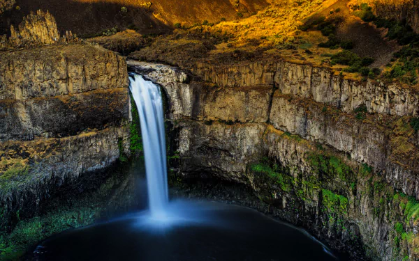 A stunning 4K Ultra HD image of Palouse Falls cascading from a cliff into a deep pool, surrounded by rugged natural rock formations and lush greenery.