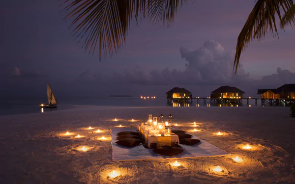 4K Ultra HD PC desktop wallpaper: candlelit beach dinner on sand at night, circular candles, overwater bungalows on horizon, sailboat under cloud-streaked sky — holiday photography background.