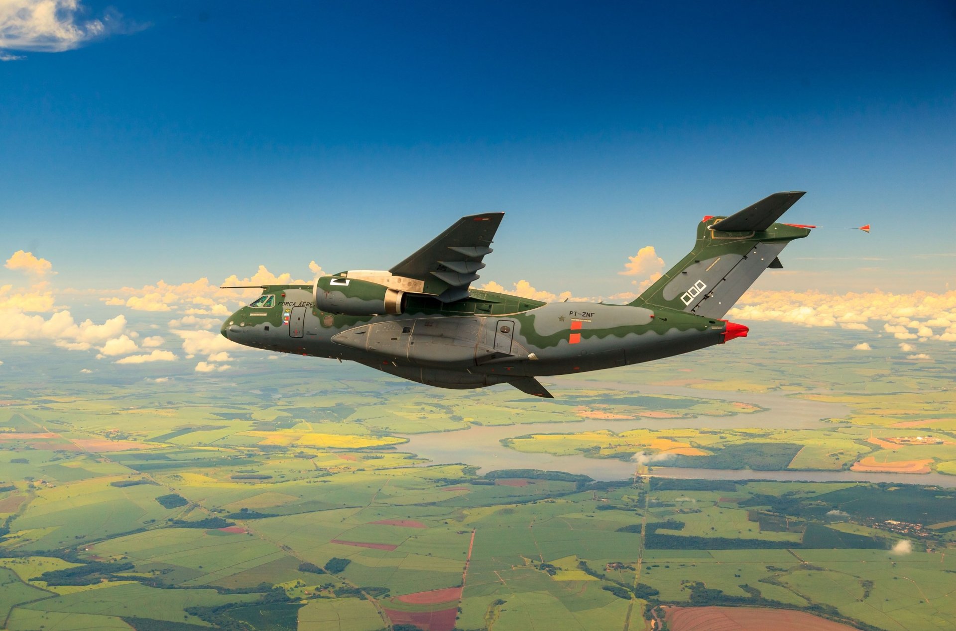 A military Embraer KC-390 transport warplane flying over a patchwork landscape under a blue sky, shown in an HD desktop wallpaper background.