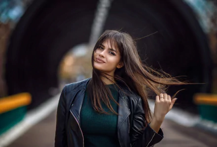 A brunette woman with brown eyes and a leather jacket smiles confidently, hair flowing, set against a blurred tunnel background with a shallow depth of field.