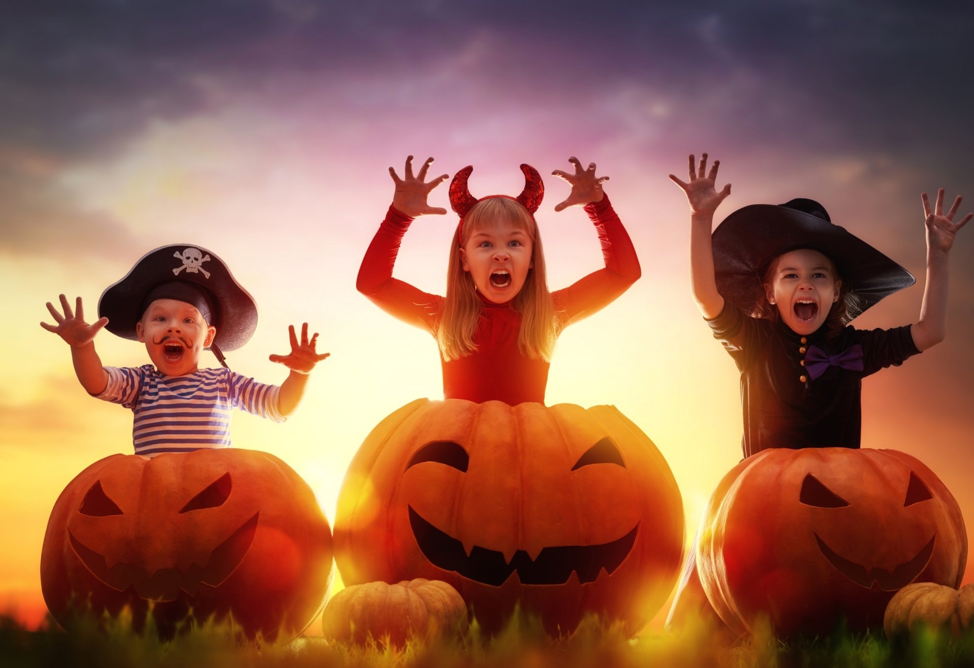 Holiday Halloween 2K Quad HD PC desktop wallpaper and background: three kids in costumes emerging from glowing jack-o'-lanterns at sunset, playful spooky scene.