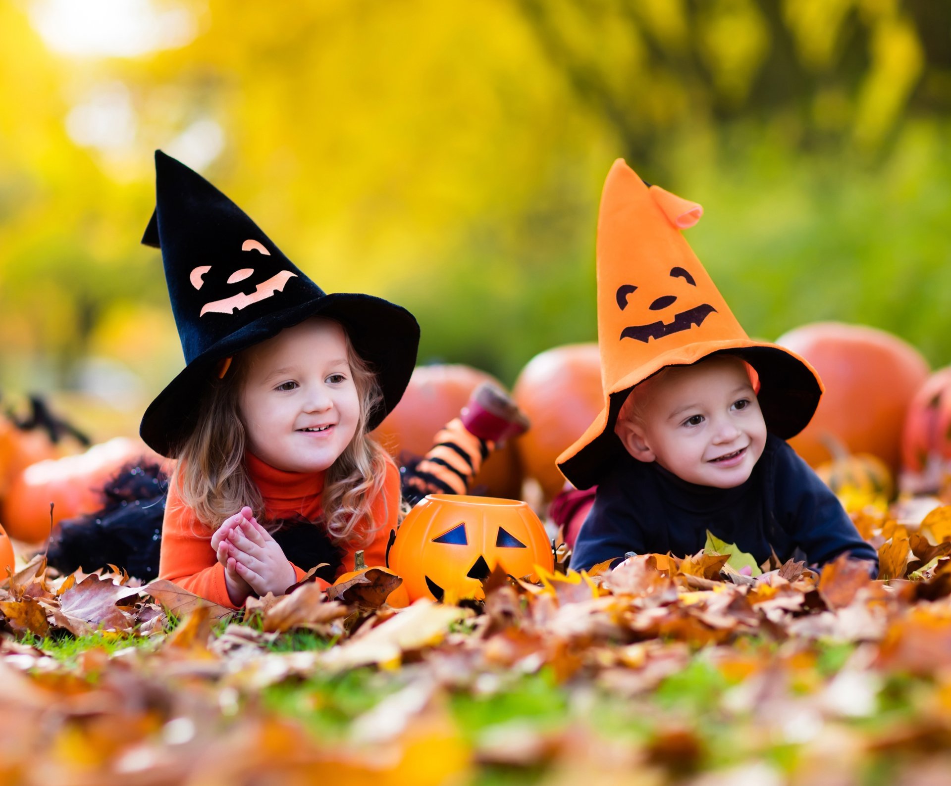 Holiday Halloween 2K Quad HD PC desktop wallpaper: two young children in orange and black witch hats smiling among pumpkins and autumn leaves.
