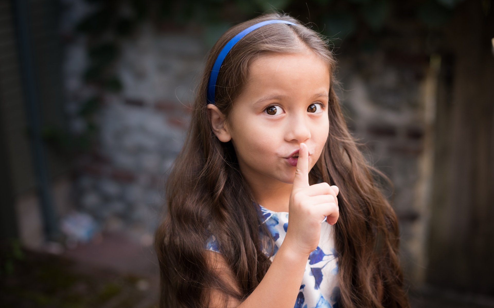 A little girl with brown eyes and long brunette hair playfully holds a finger to her lips, asking for silence, captured in a charming 4K Ultra HD image.