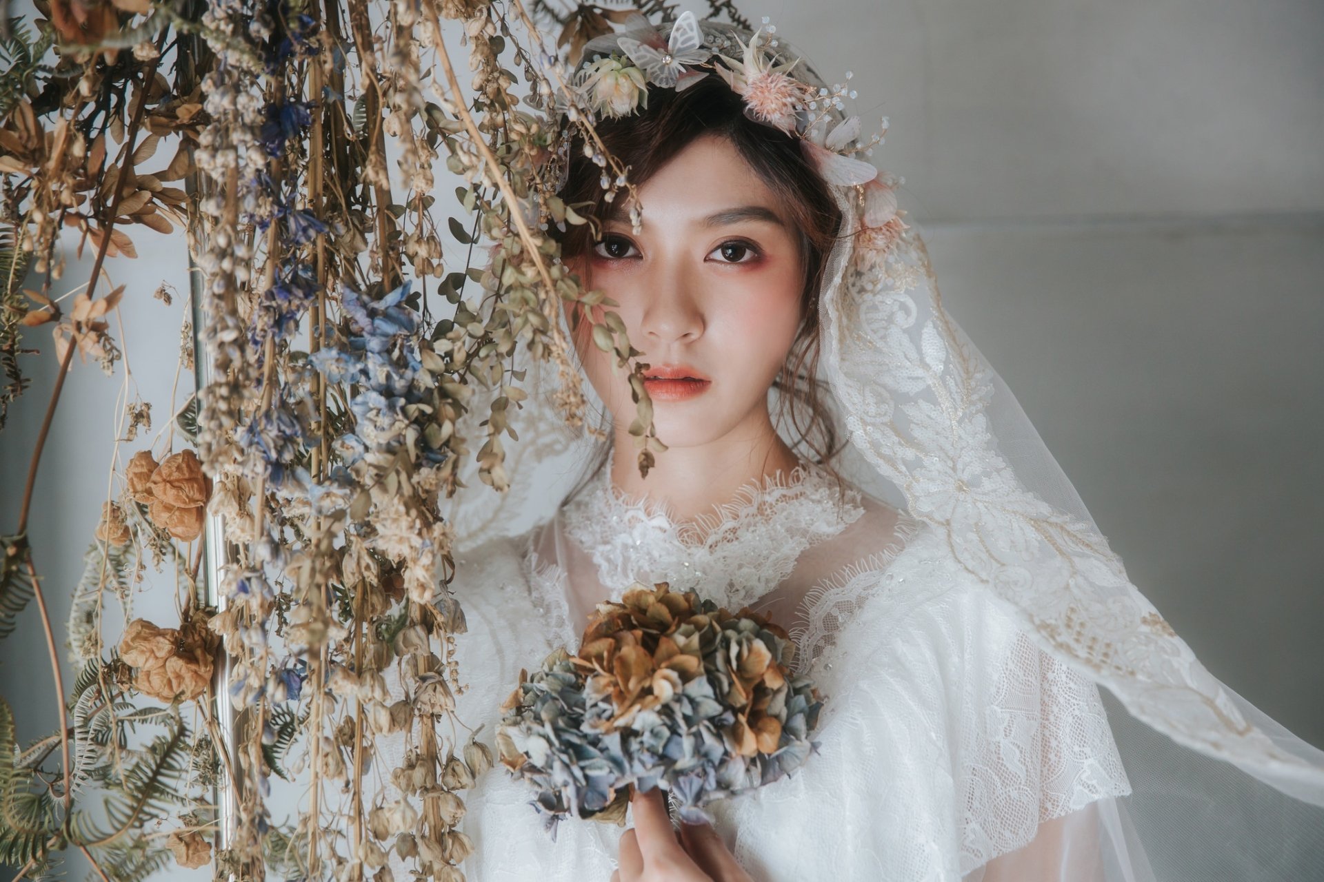 Brunette bride with brown eyes in a lace wedding dress and veil holds a dried-flower bouquet beside hanging blooms — elegant bridal model HD desktop wallpaper.