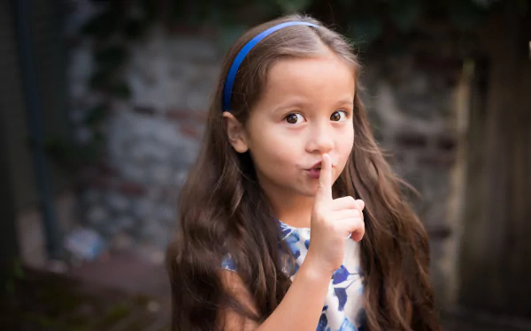 A little girl with brown eyes and long brunette hair playfully holds a finger to her lips, asking for silence, captured in a charming 4K Ultra HD image.