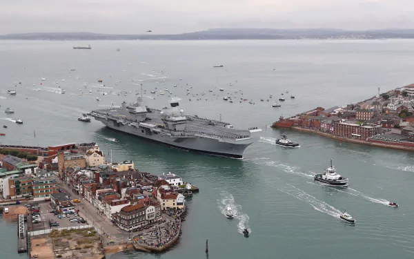 HD desktop wallpaper showing the HMS Queen Elizabeth (R08) aircraft carrier, a prominent military warship, navigating through a busy coastal harbor.
