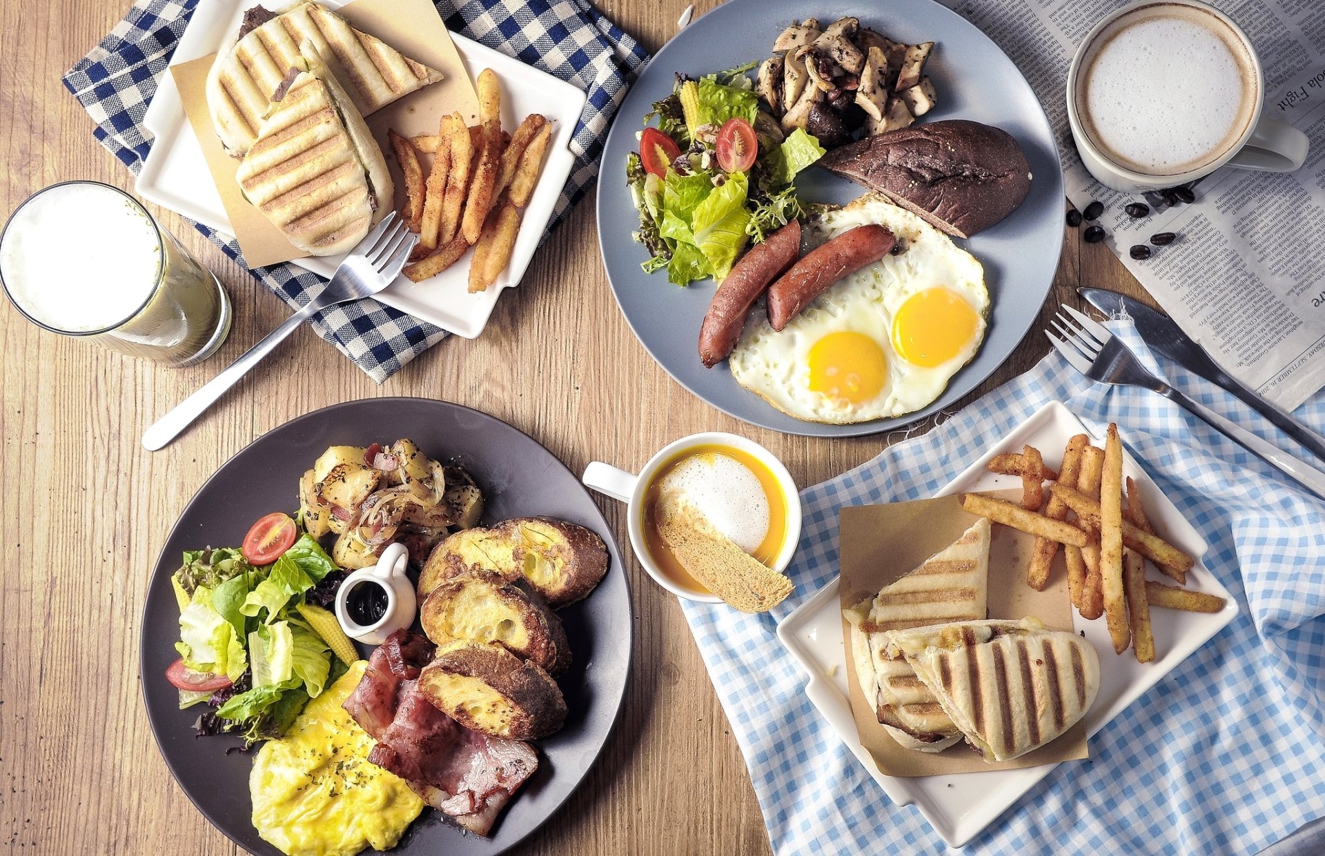 HD desktop wallpaper of a breakfast still life featuring eggs, sausage, salad, fries, toast, and a cup of milk on a wooden table.
