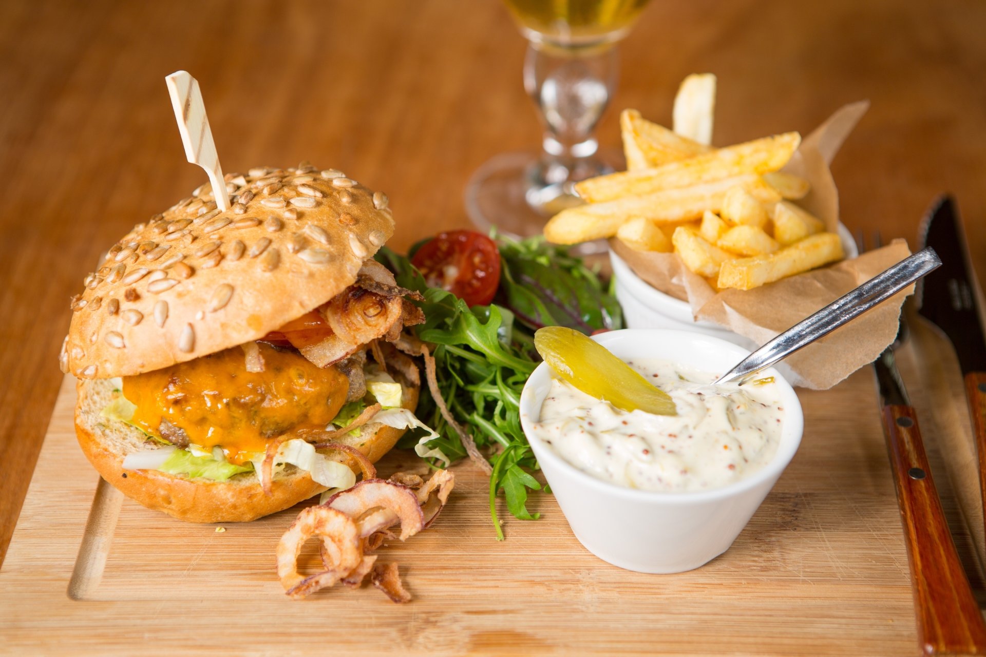 HD desktop wallpaper featuring a juicy burger with melted cheese, crispy French fries, fresh salad, and a creamy dipping sauce on a wooden board.