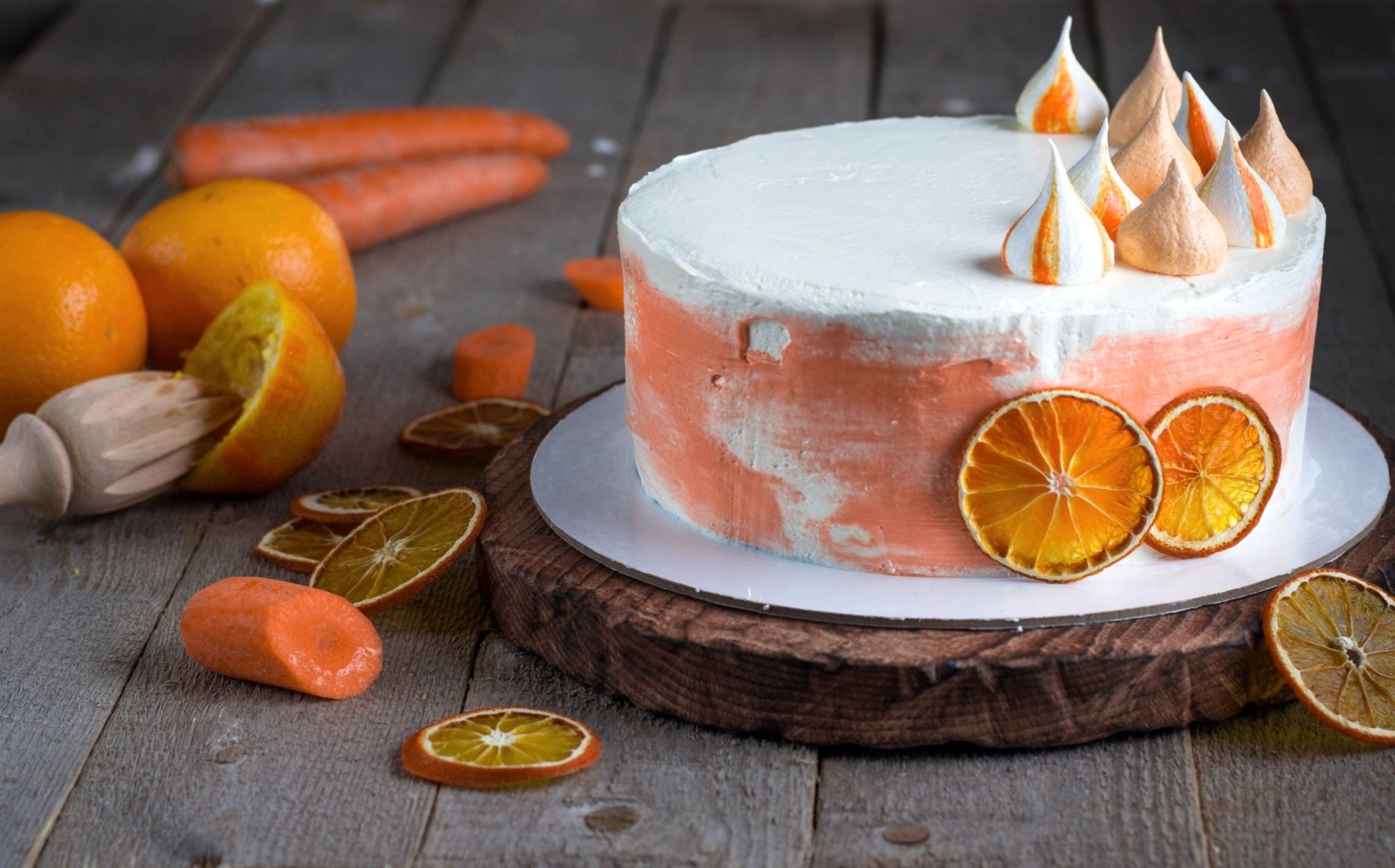 A still life HD desktop wallpaper featuring a carrot cake with frosted peaks, surrounded by fresh carrots and sliced oranges on a rustic wooden surface.