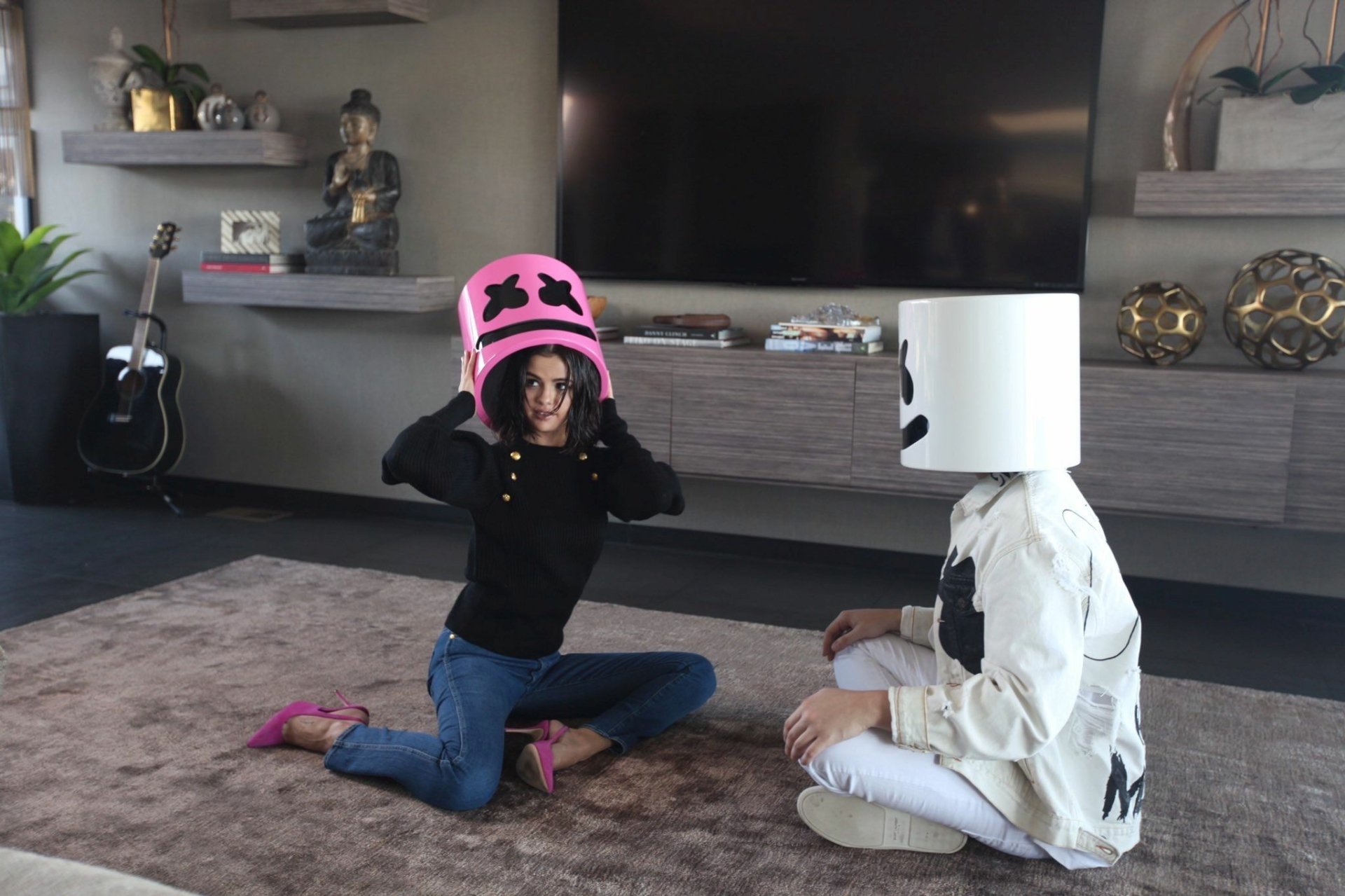 A fun moment featuring Marshmello and Selena Gomez, both wearing iconic marshmallow helmets, sitting on a rug in a stylish interior. A vibrant music-themed wallpaper for fans.