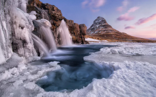 Winter scene of Kirkjufoss waterfall cascading over icy rocks with snow-covered Kirkjufell mountain in the background, Iceland. HD nature desktop wallpaper.