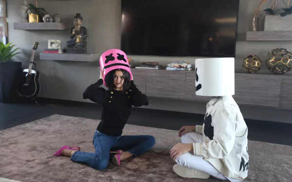 A fun moment featuring Marshmello and Selena Gomez, both wearing iconic marshmallow helmets, sitting on a rug in a stylish interior. A vibrant music-themed wallpaper for fans.