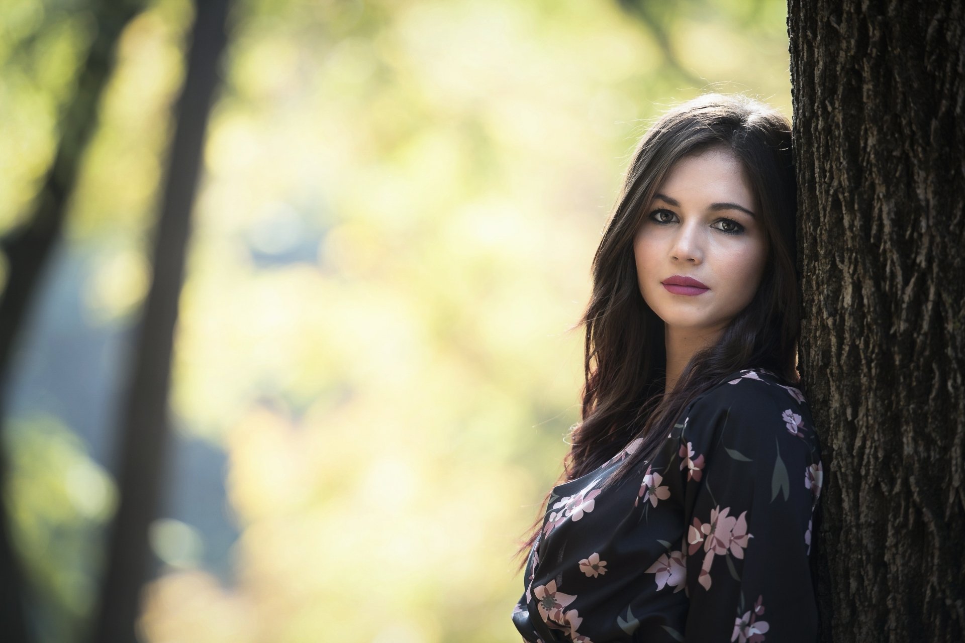 HD desktop wallpaper of a brunette woman with lipstick, posing near a tree with a soft depth of field, creating a serene and elegant background.