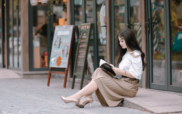A brunette Asian woman in high heels sits reading a book on a city sidewalk, creating a serene HD desktop wallpaper.