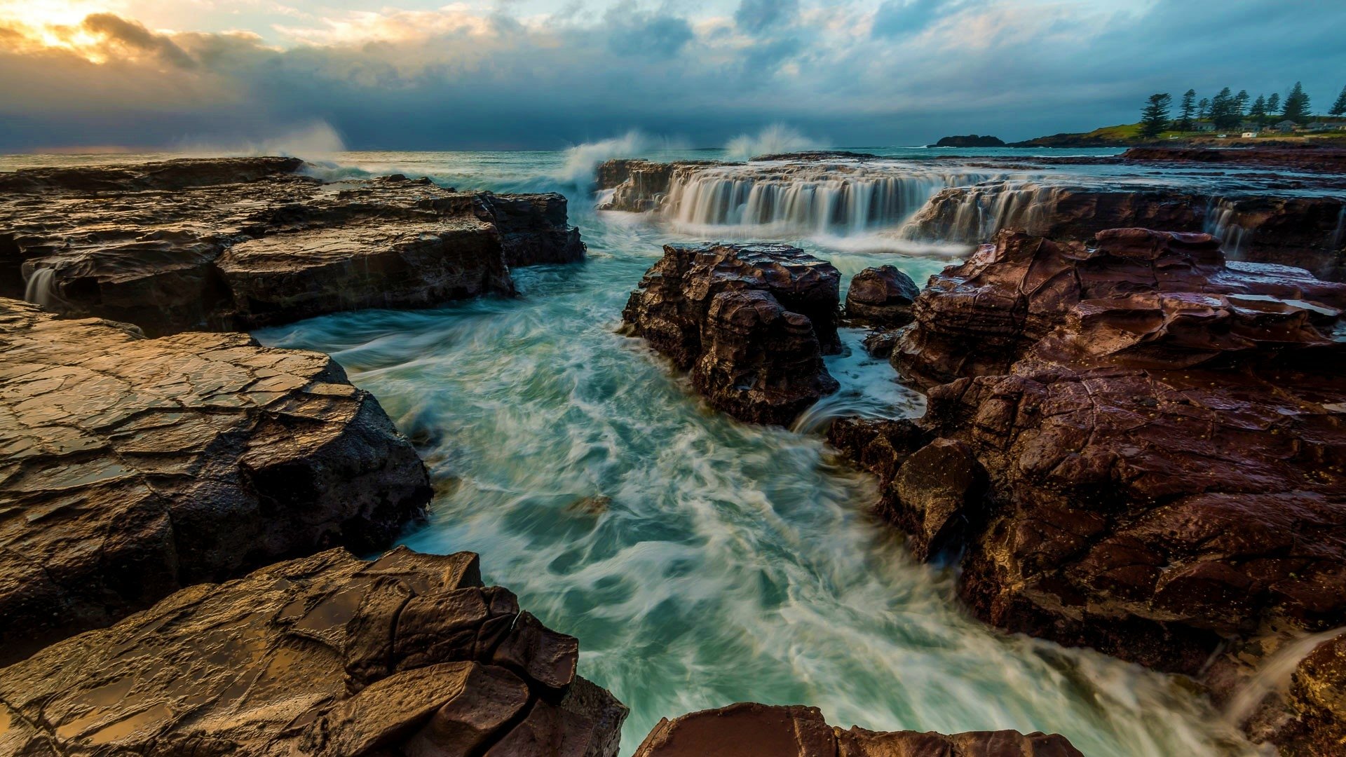 HD desktop wallpaper showing a stunning natural scene of ocean waves crashing against rocky formations under a cloudy sky on the horizon. The image captures the dynamic beauty of the sea and nature.