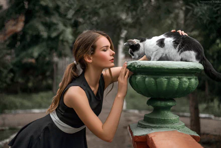 A woman in a black dress and ponytail interacts affectionately with a cat on a green fountain, set against a softly blurred natural background.