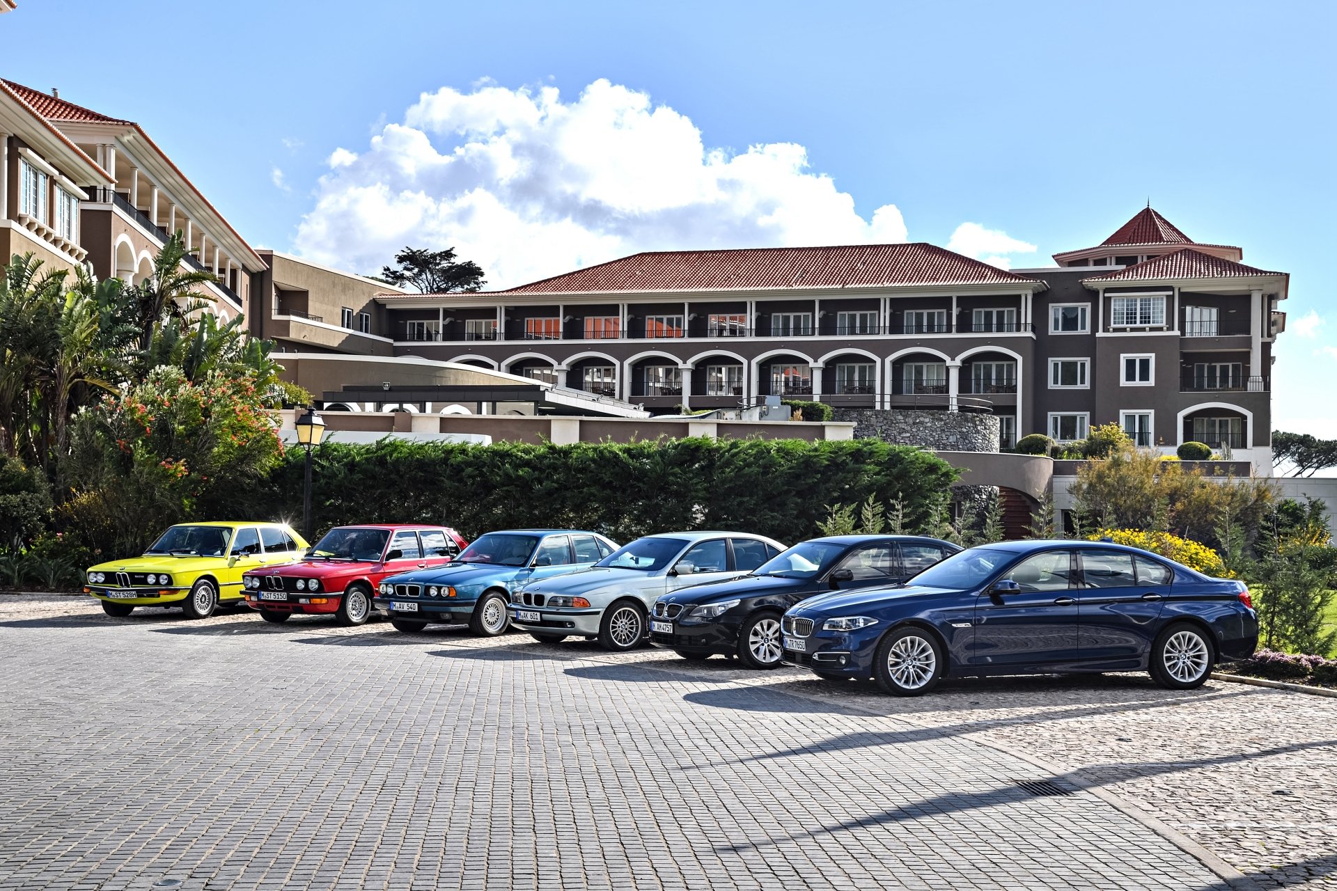 A lineup of colorful BMW cars, including a standout yellow vehicle, parked in front of a large, elegant building, captured in 4K Ultra HD.