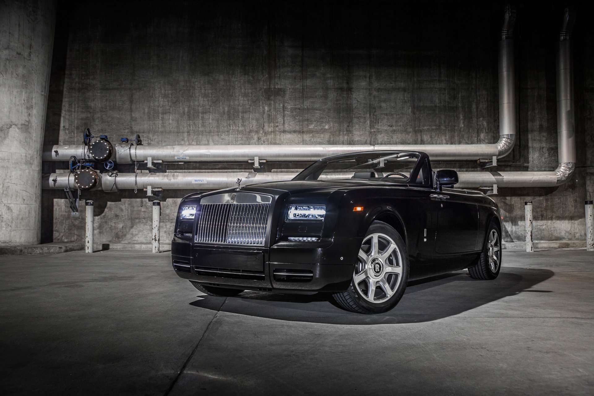 HD PC wallpaper featuring a sleek black Rolls-Royce Phantom convertible parked in an industrial setting with exposed pipes and concrete walls.