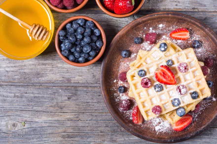 A 4K Ultra HD image of a waffle topped with raspberry, strawberry, and blueberry, dusted with powdered sugar, accompanied by a bowl of honey and a bowl of fresh blueberries.