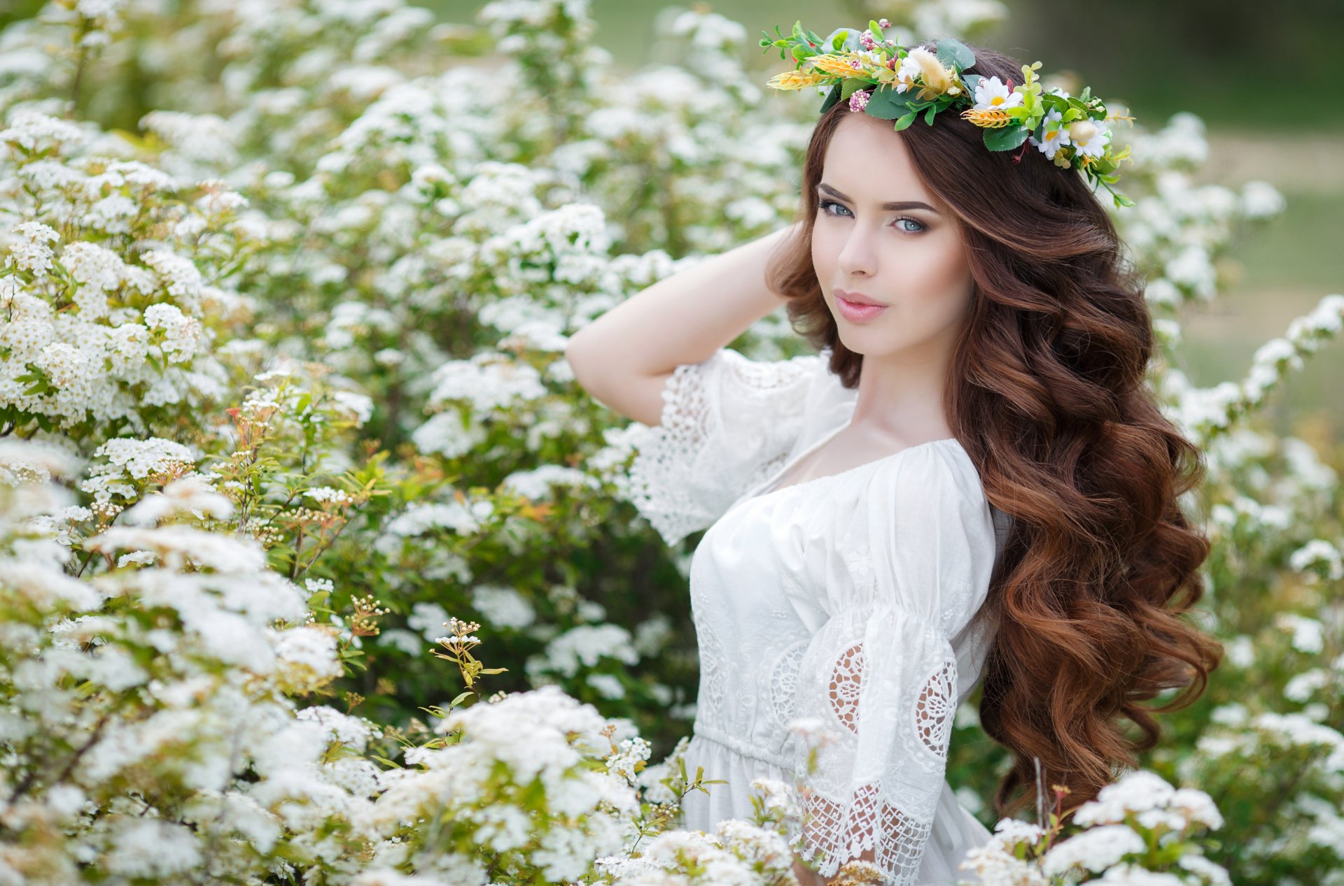 A woman with long brunette hair and blue eyes, wearing a floral wreath, poses among white flowers, suitable as a HD desktop wallpaper.
