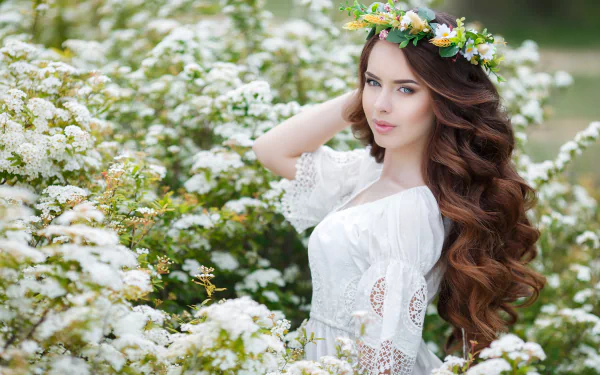 A woman with long brunette hair and blue eyes, wearing a floral wreath, poses among white flowers, suitable as a HD desktop wallpaper.