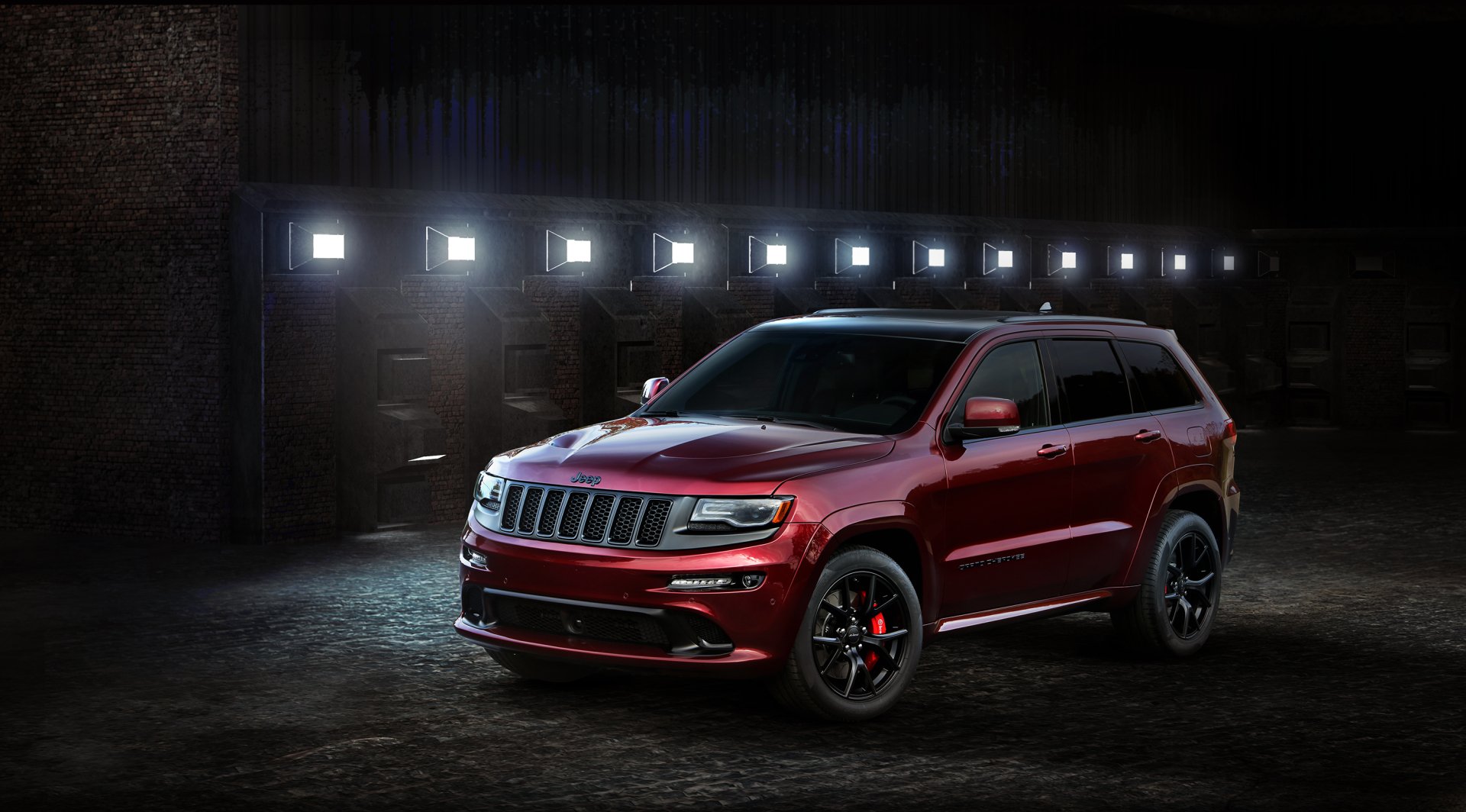 Red Jeep Grand Cherokee vehicle parked in a dim industrial garage, lit by a row of wall lights — 2K Quad HD PC desktop wallpaper/background.