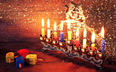 HD desktop wallpaper featuring a glowing Hanukkah menorah with lit candles and colorful dreidels on a wooden surface, surrounded by festive golden sparkles.