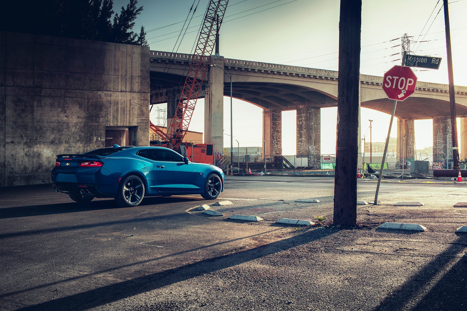 HD desktop wallpaper featuring a blue Chevrolet Camaro parked under an urban overpass with a stop sign and construction cranes in the background.