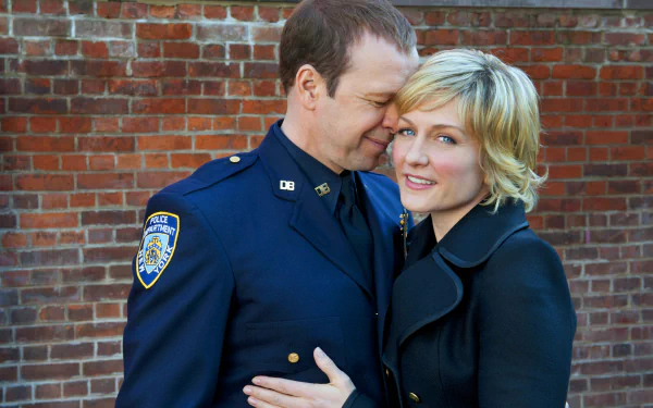 HD desktop wallpaper of Blue Bloods featuring a smiling police officer in uniform embracing a woman with a short haircut against a brick wall background.