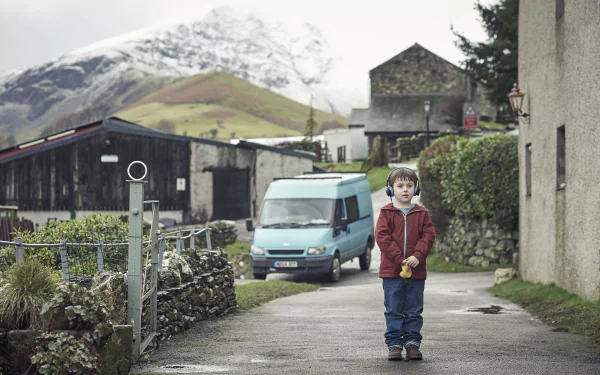 HD desktop wallpaper of The A Word, featuring a young boy standing on a village road with mountains in the background.