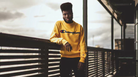HD desktop wallpaper of a man in a yellow Champion sweatshirt leaning on a railing, with moody skies in the background.