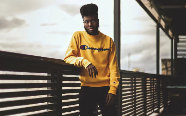 HD desktop wallpaper of a man in a yellow Champion sweatshirt leaning on a railing, with moody skies in the background.