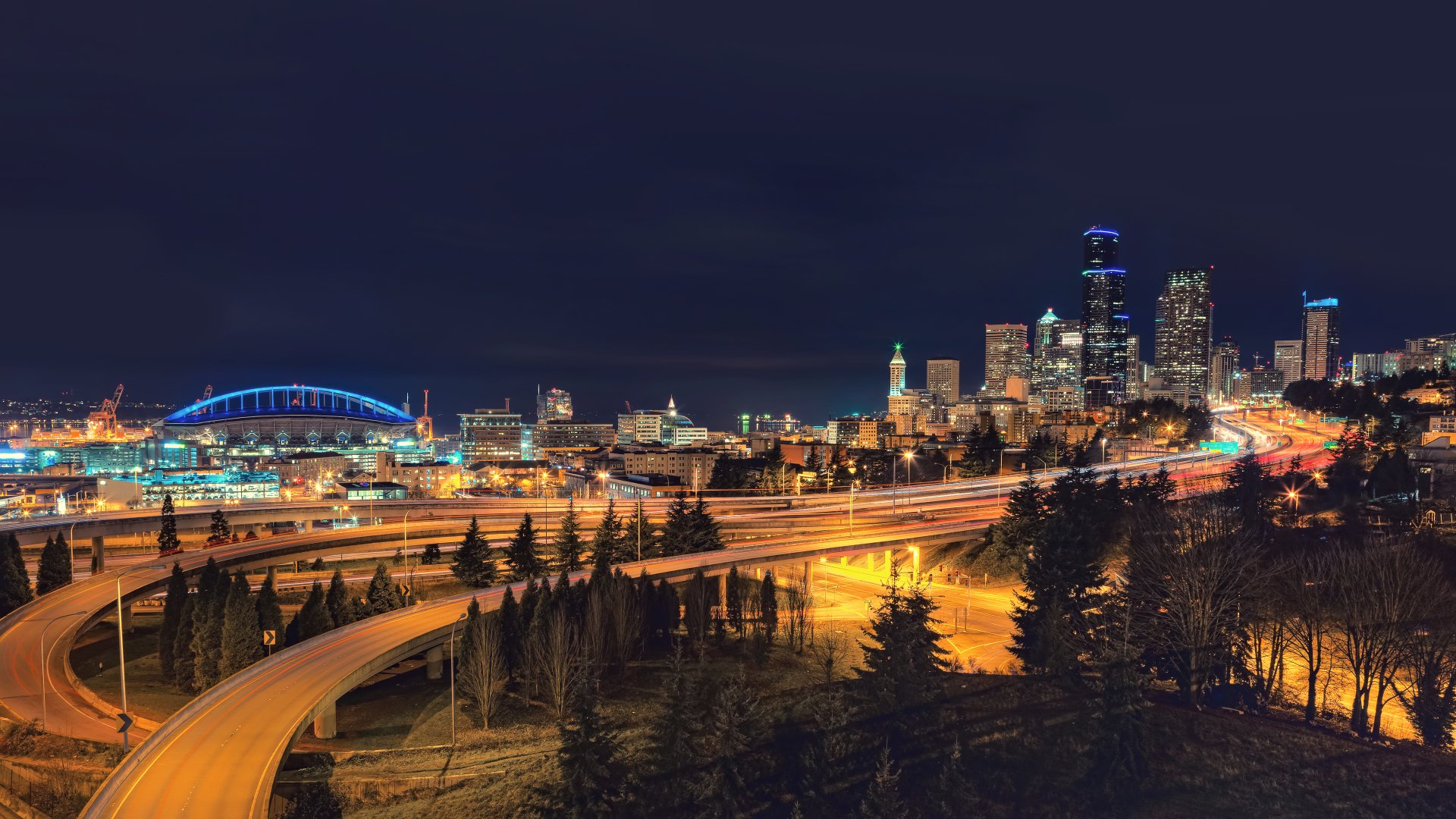 4K Ultra HD nighttime cityscape of Seattle featuring illuminated freeways winding through the urban skyline with prominent buildings and structures under a dark sky.