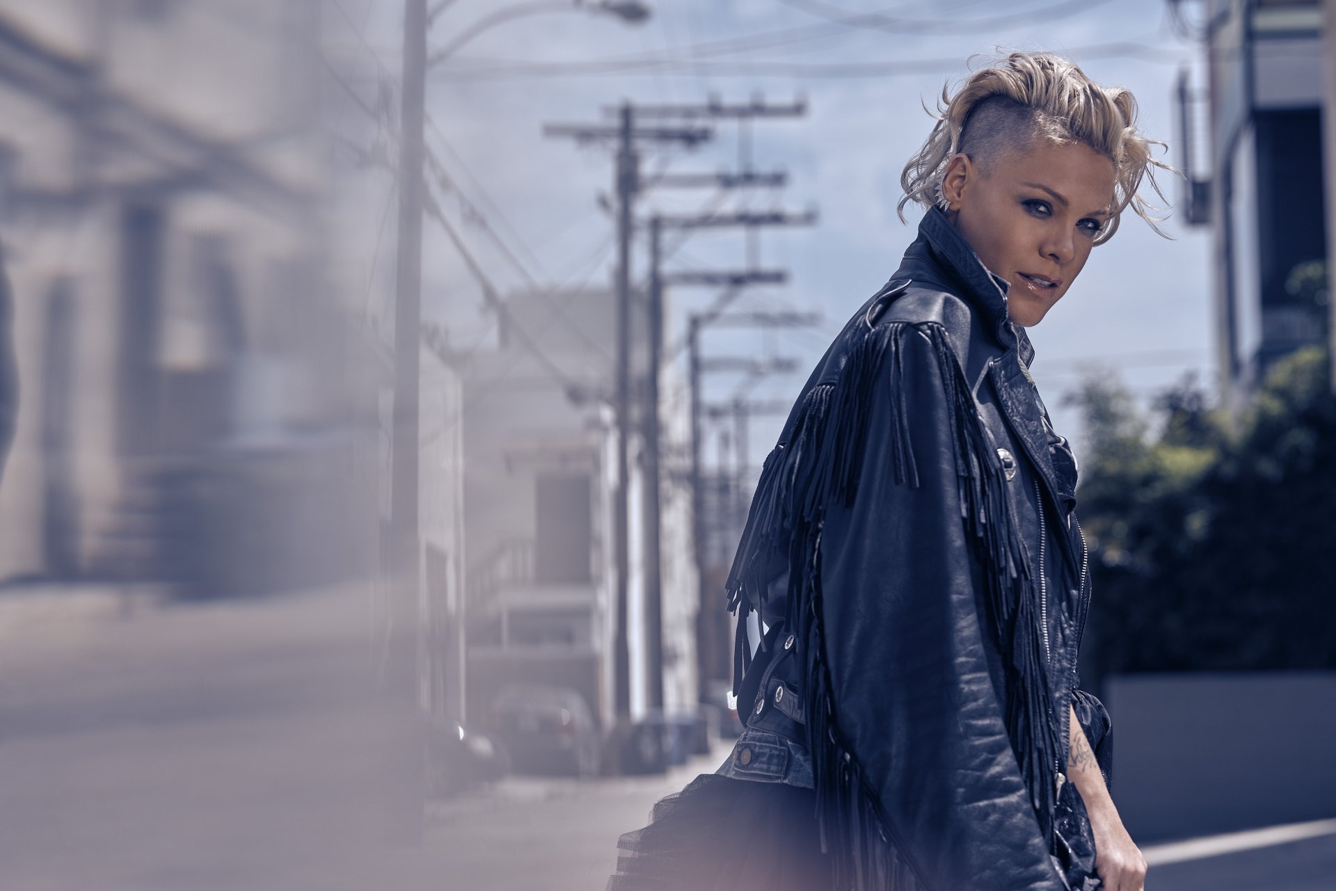Blonde singer Pink with short hair, wearing a black leather jacket, captured in 4K Ultra HD with shallow depth of field against an urban street backdrop.