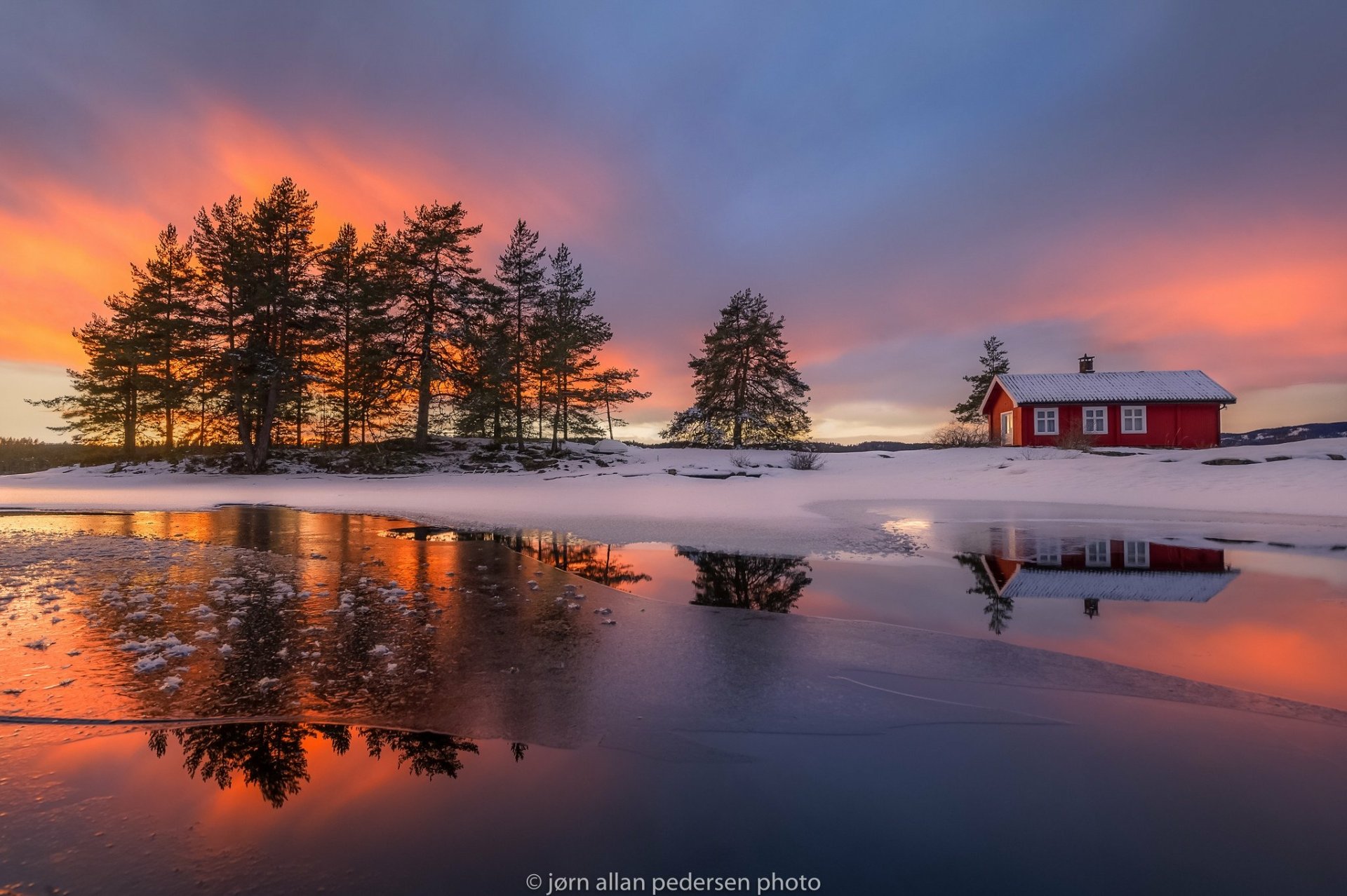 HD desktop wallpaper featuring a winter scene with a house near a frozen lake, snow-covered ground, trees, and a stunning sunset reflecting on the icy water.