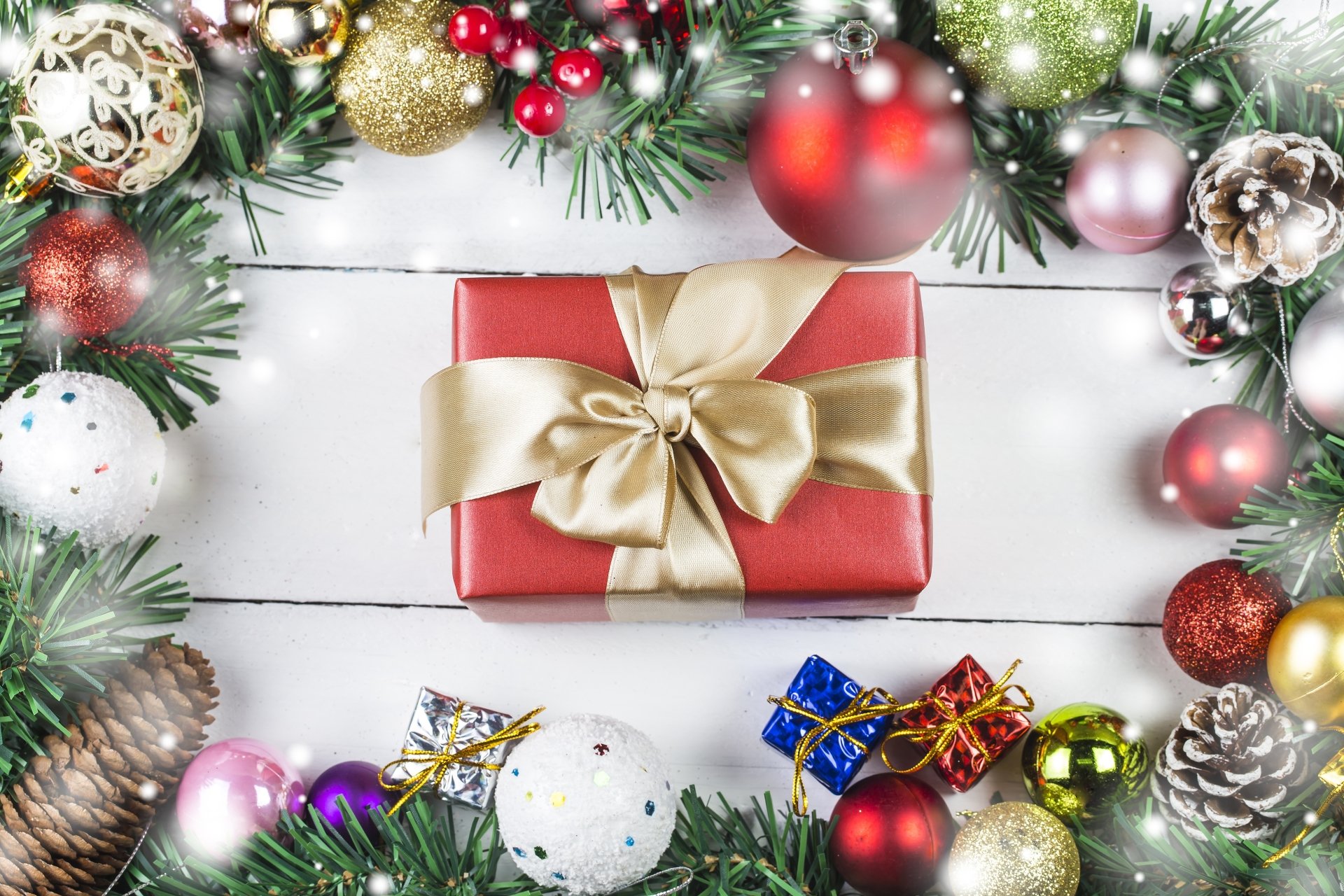 A festive 4K Ultra HD Christmas wallpaper featuring a red gift box with a gold bow, surrounded by holiday baubles, ornaments, and greenery on a white wooden background.