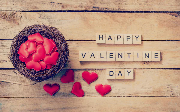 Romantic HD desktop wallpaper featuring a nest filled with heart-shaped objects and scrabble tiles spelling Happy Valentine's Day on a wooden background, celebrating love and the holiday.