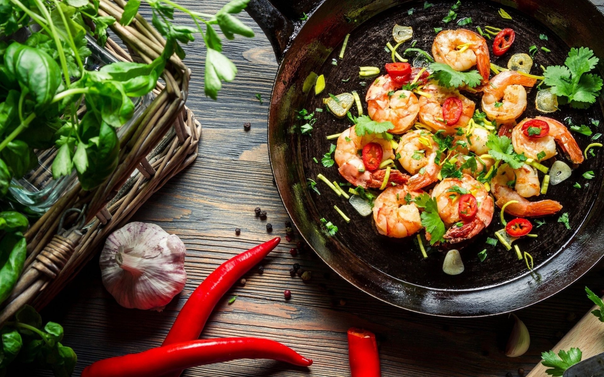 HD desktop wallpaper showing a still life of cooked shrimp with sliced red peppers and herbs on a black skillet, surrounded by fresh garlic, chili, and greens.