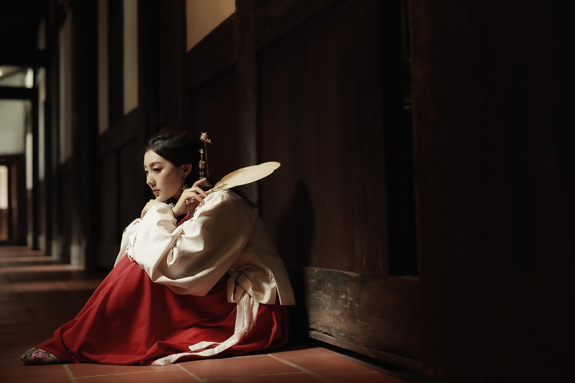 An Asian brunette woman in traditional costume sits on the floor of a dimly lit wooden hallway, holding a fan, captured in an HD desktop wallpaper style.