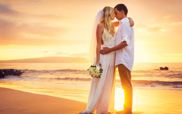 Romantic wedding moment of a bride in a wedding dress and groom embracing on a beach at sunset, captured in stunning HD photography for a PC desktop wallpaper.