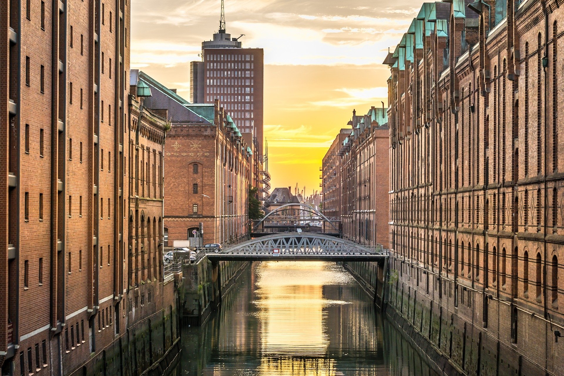 HD desktop wallpaper of Hamburg, Germany: brick warehouse buildings flank a canal with an iron bridge spanning the water at sunset, a striking man-made urban scene.