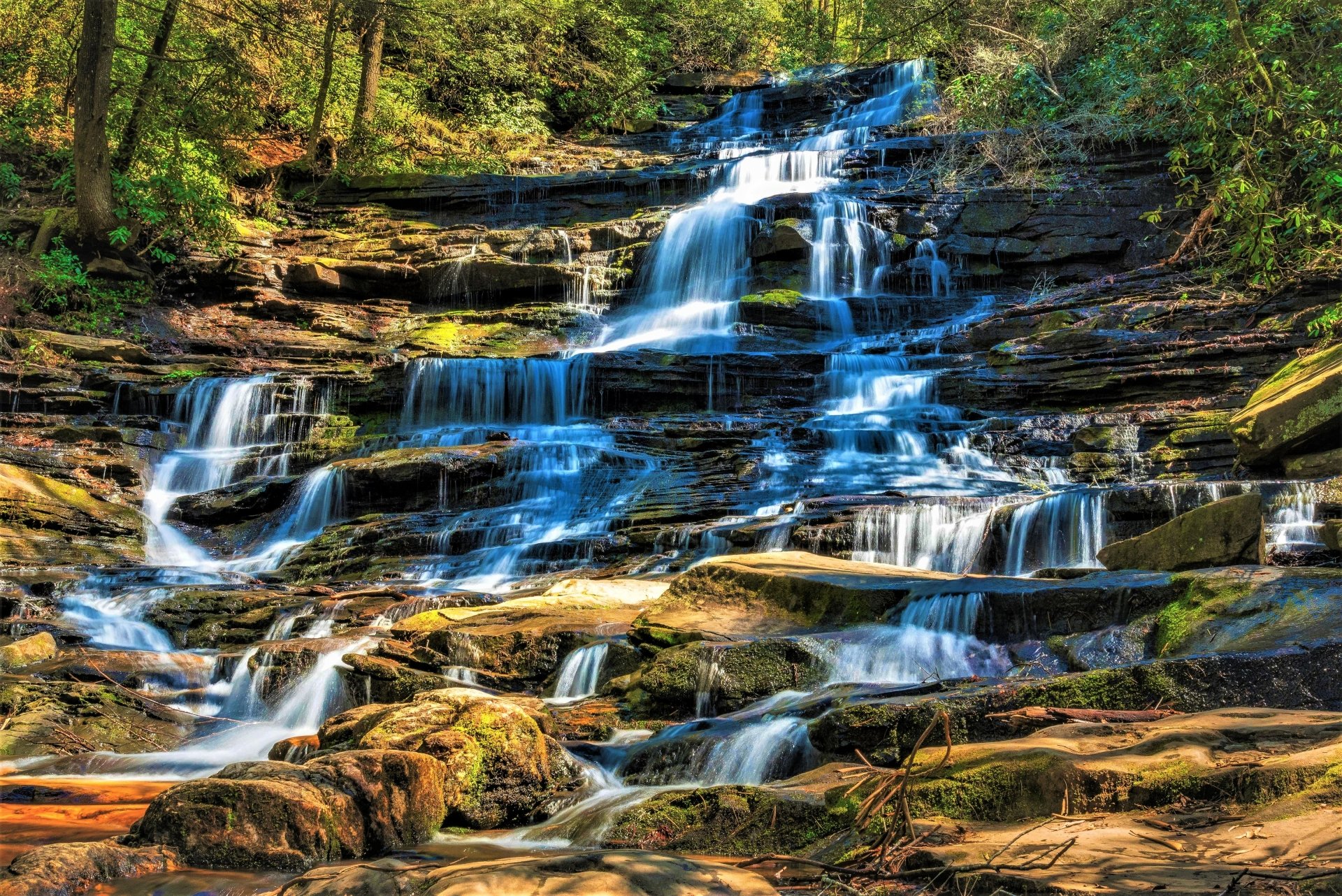 4K Ultra HD image of a cascading waterfall flowing over layered stones surrounded by lush green nature in a serene forest setting.