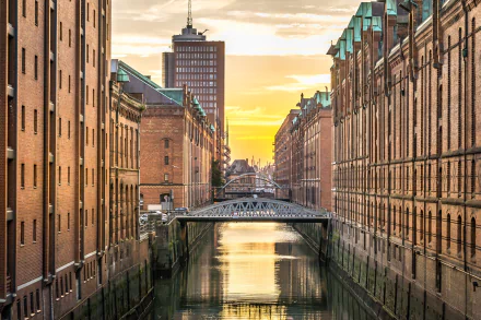 HD desktop wallpaper of Hamburg, Germany: brick warehouse buildings flank a canal with an iron bridge spanning the water at sunset, a striking man-made urban scene.
